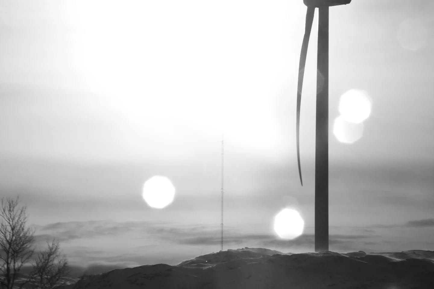 Black and white photo of a wind turbine and thin radio tower on a hill, with lens flares and hazy sky.