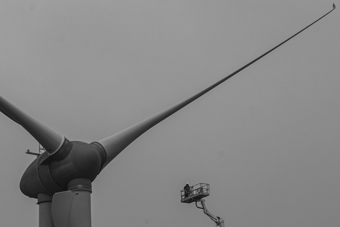 Black-and-white photo of a wind turbine hub and blades against a gray sky, with a worker on a crane platform nearby.