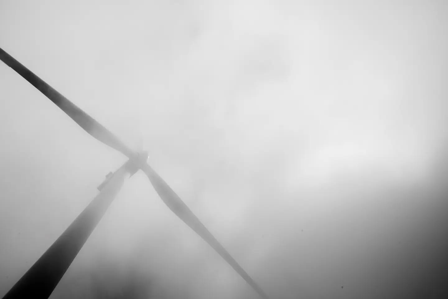 Black and white photo of wind turbine blades against a foggy, hazy sky, viewed from below.