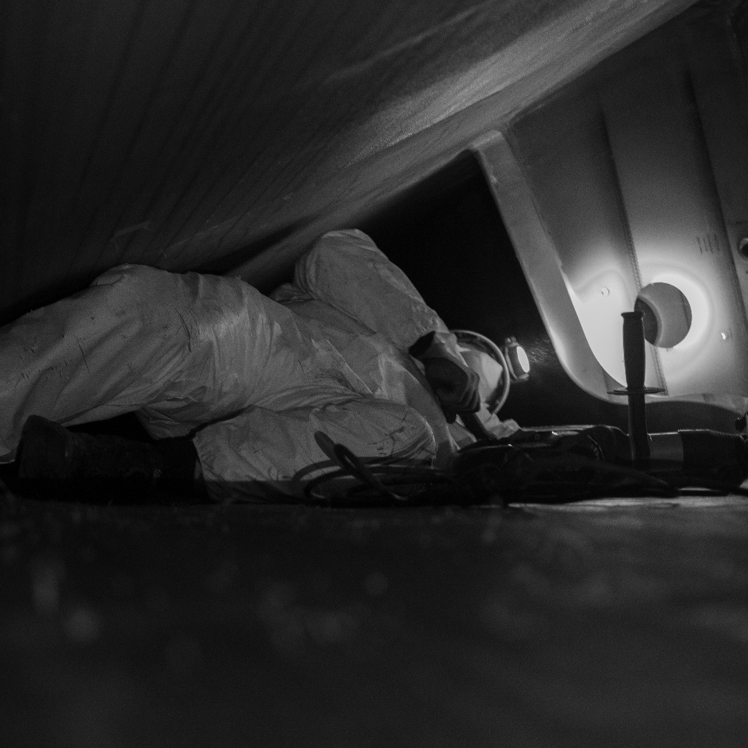 Black and white photo of a person in coveralls crawling through a tight crawl space, using a flashlight, tools nearby.