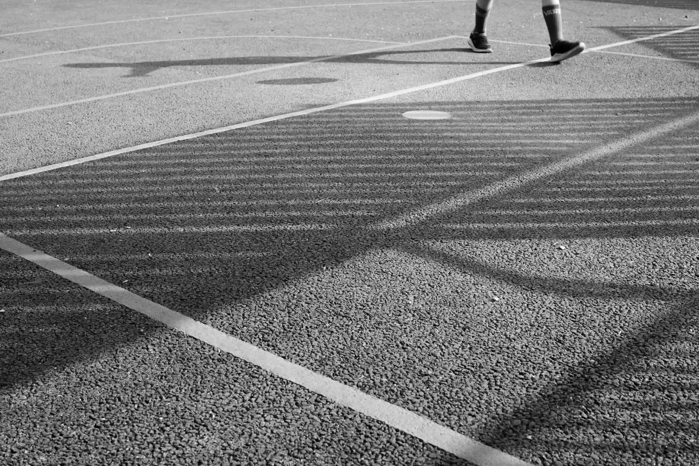 Black and white photo of a person's legs and sneakers on a court, with long fence shadows crossing painted lines on the pavement.