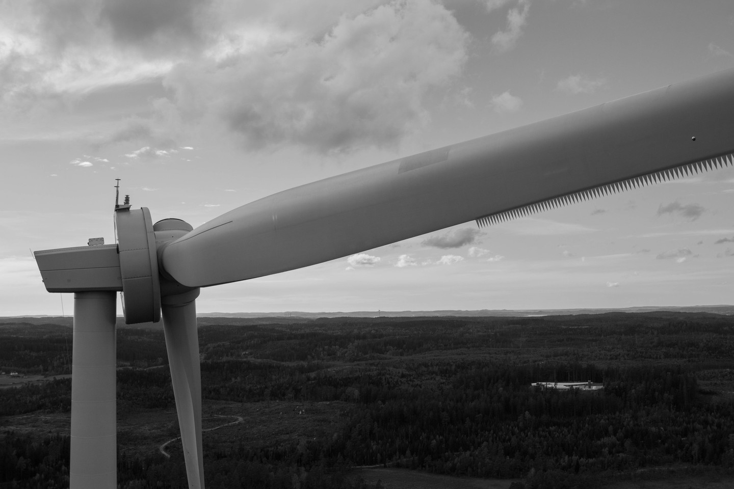Black and white close-up of a wind turbine nacelle and blades against a cloudy sky, overlooking a forested landscape below.