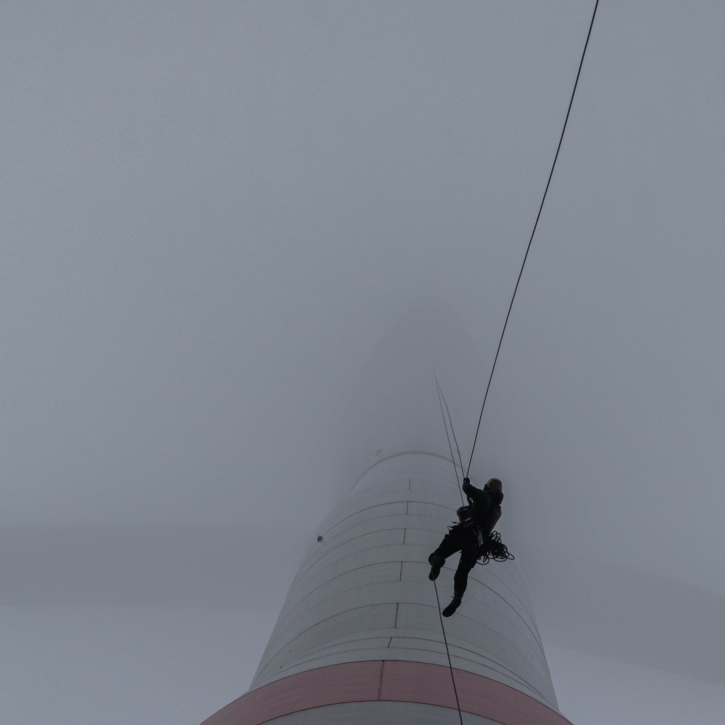 Silhouette of a rope-access technician rappelling down a tall structure, disappearing into thick fog above.