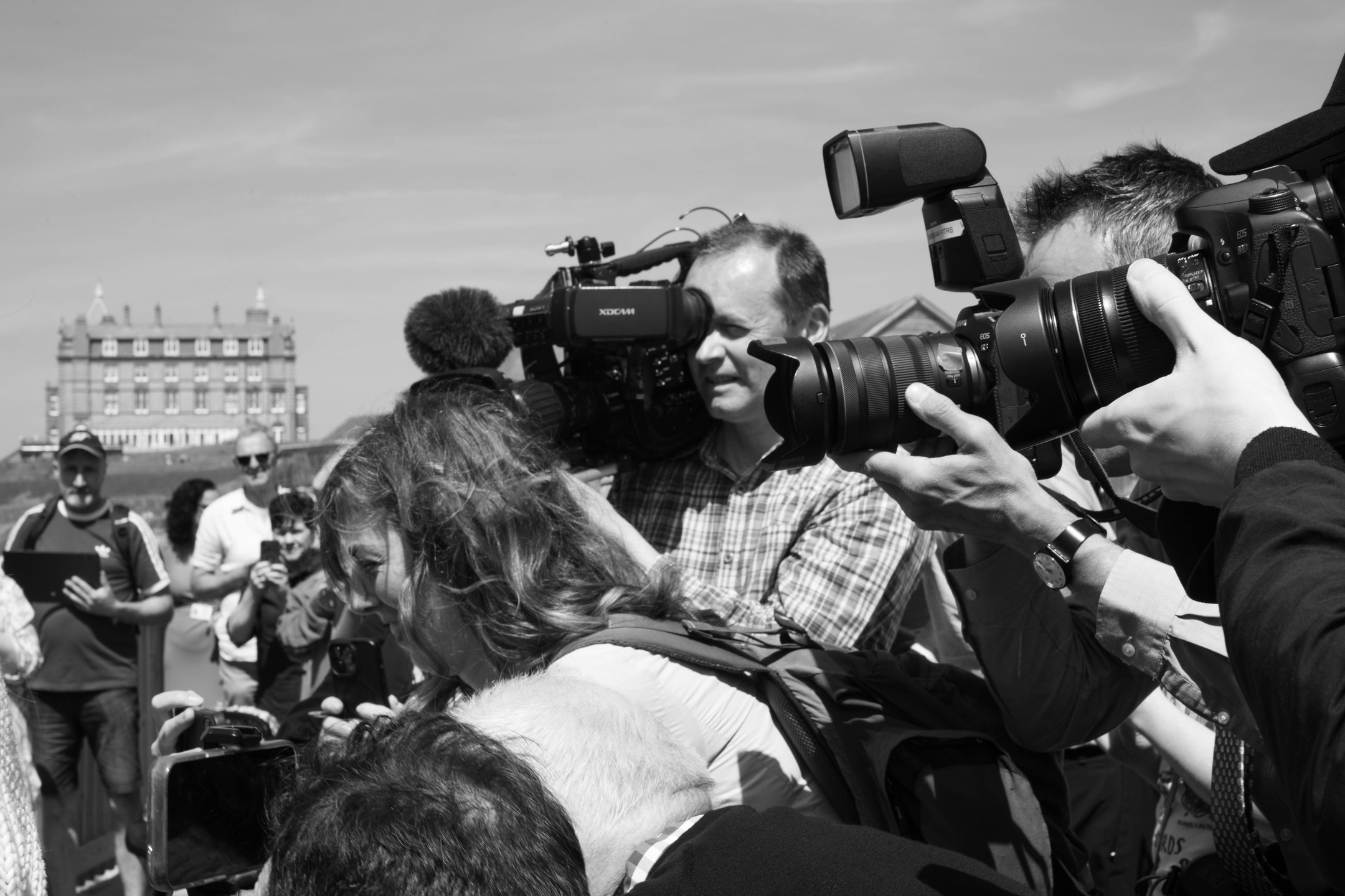 Black-and-white photo of press photographers and a videographer crowded together with cameras raised, a large building visible in the background.