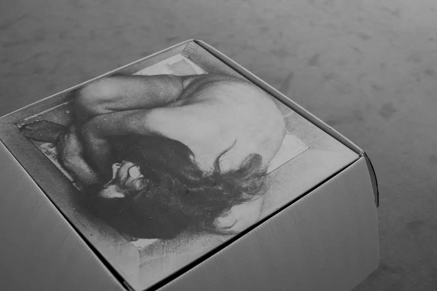 Black-and-white photo of a cardboard box with a printed image of a nude, bearded figure on its top surface, set on a gray floor.