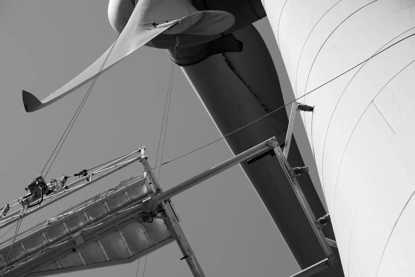 Black-and-white low-angle view of a wind turbine blade and tower with a metal maintenance platform suspended below.