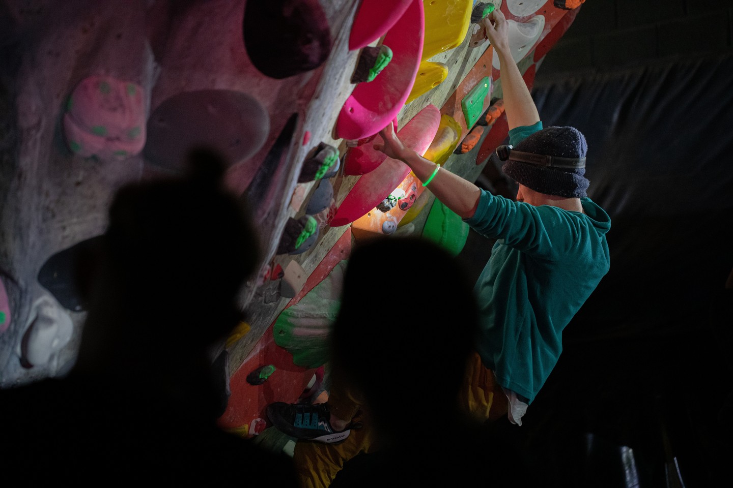 Climber in a beanie and teal top reaching for holds on a colorful bouldering wall, viewed past silhouetted onlookers.