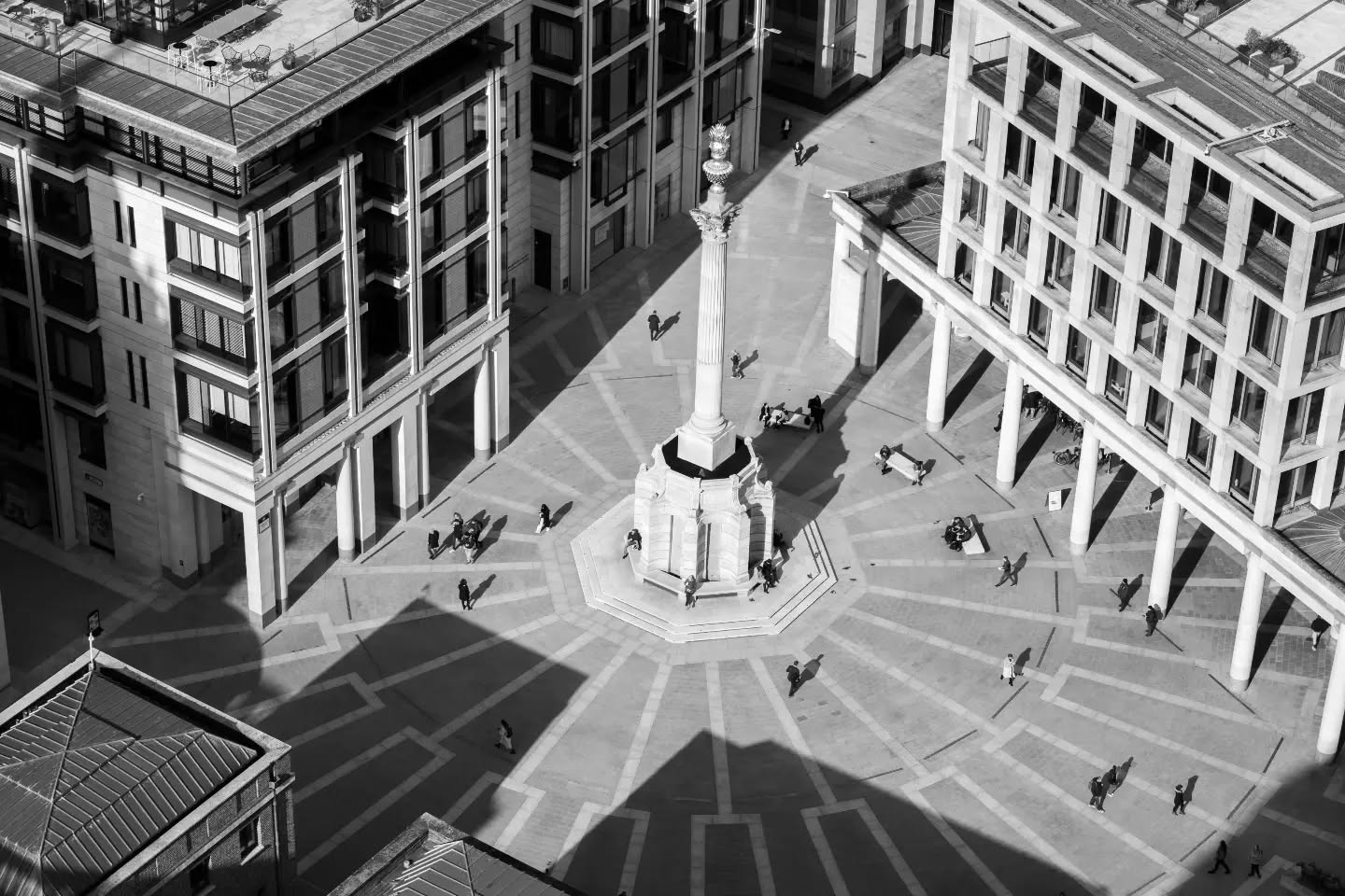 Aerial black and white view of a paved plaza with a radial pattern, a tall column at center, surrounded by buildings and scat