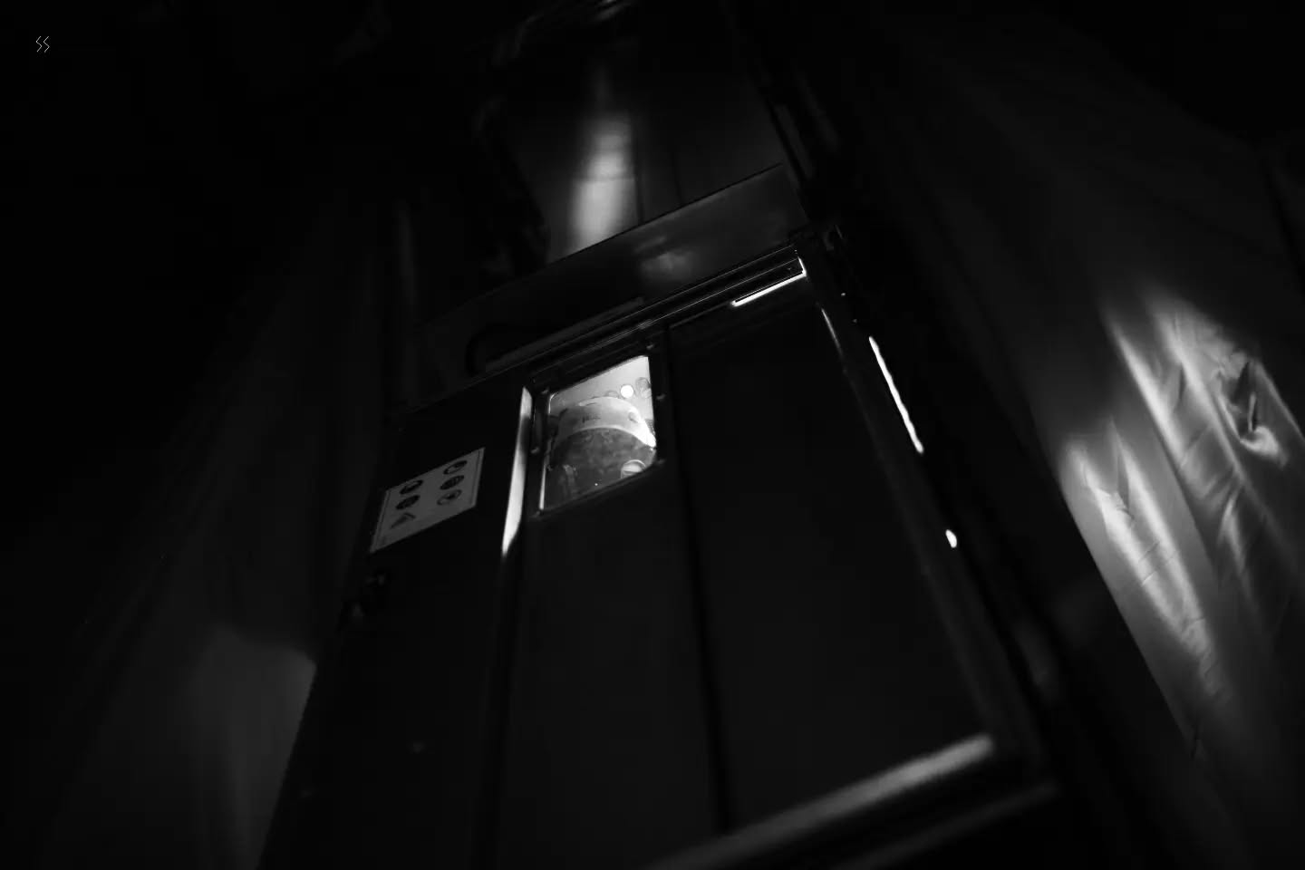 Black and white photo of a metal door with a small window and control panel, viewed at an upward angle in dim light.