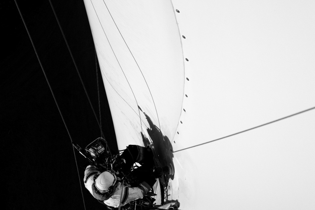 Black and white photo of a sailboat mast top with rigging wires and a crew member in a harness working aloft.