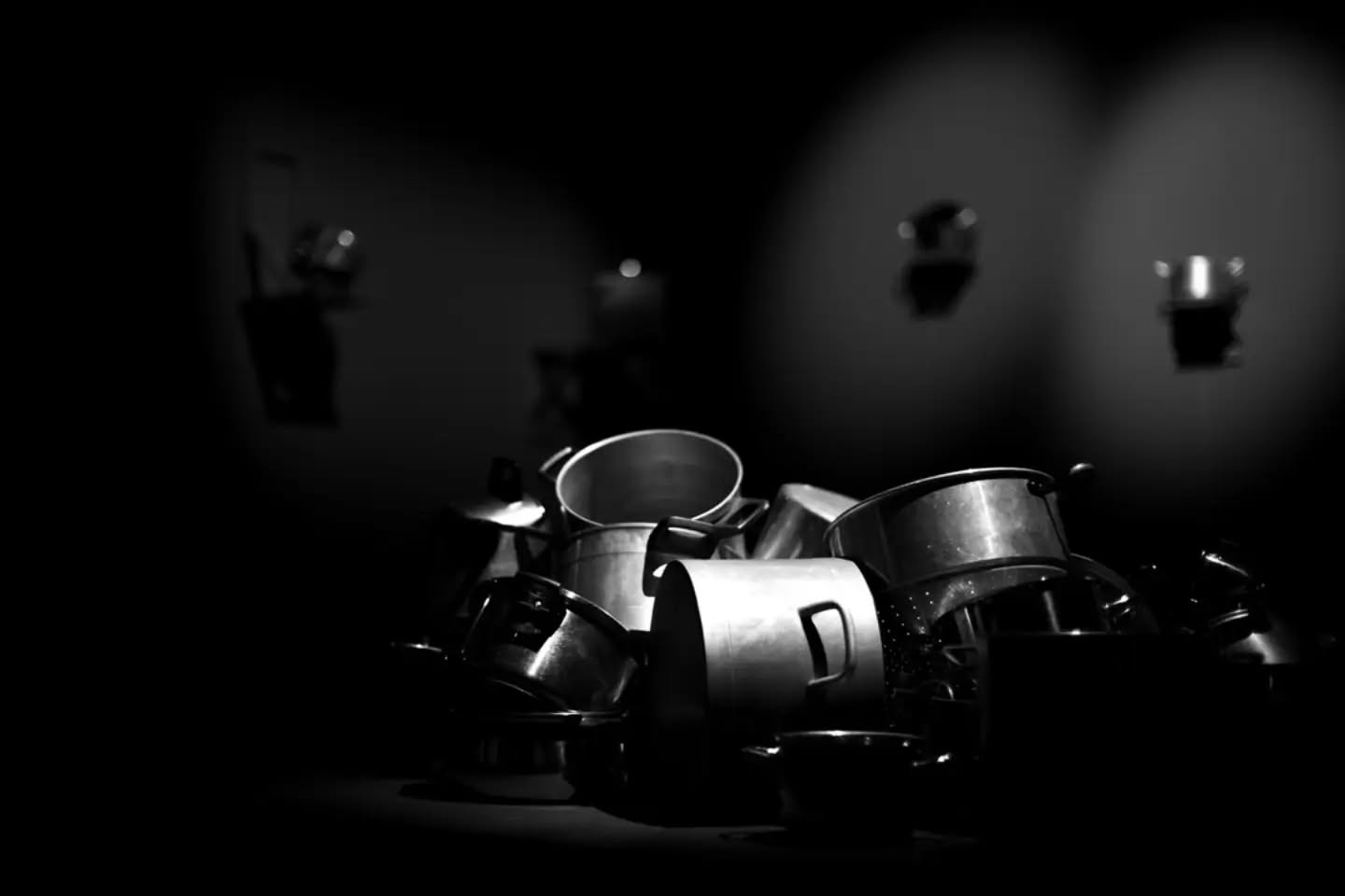 Black and white photo of a jumbled pile of metal pots and pans on a dark surface, with blurred pans hanging on a wall behind.
