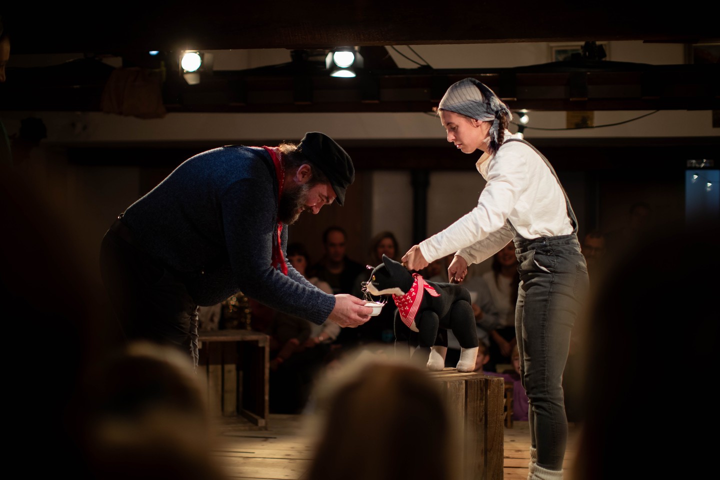 Man in cap holding a bowl kneels toward a woman controlling a black cat puppet on stage, audience seated around.
