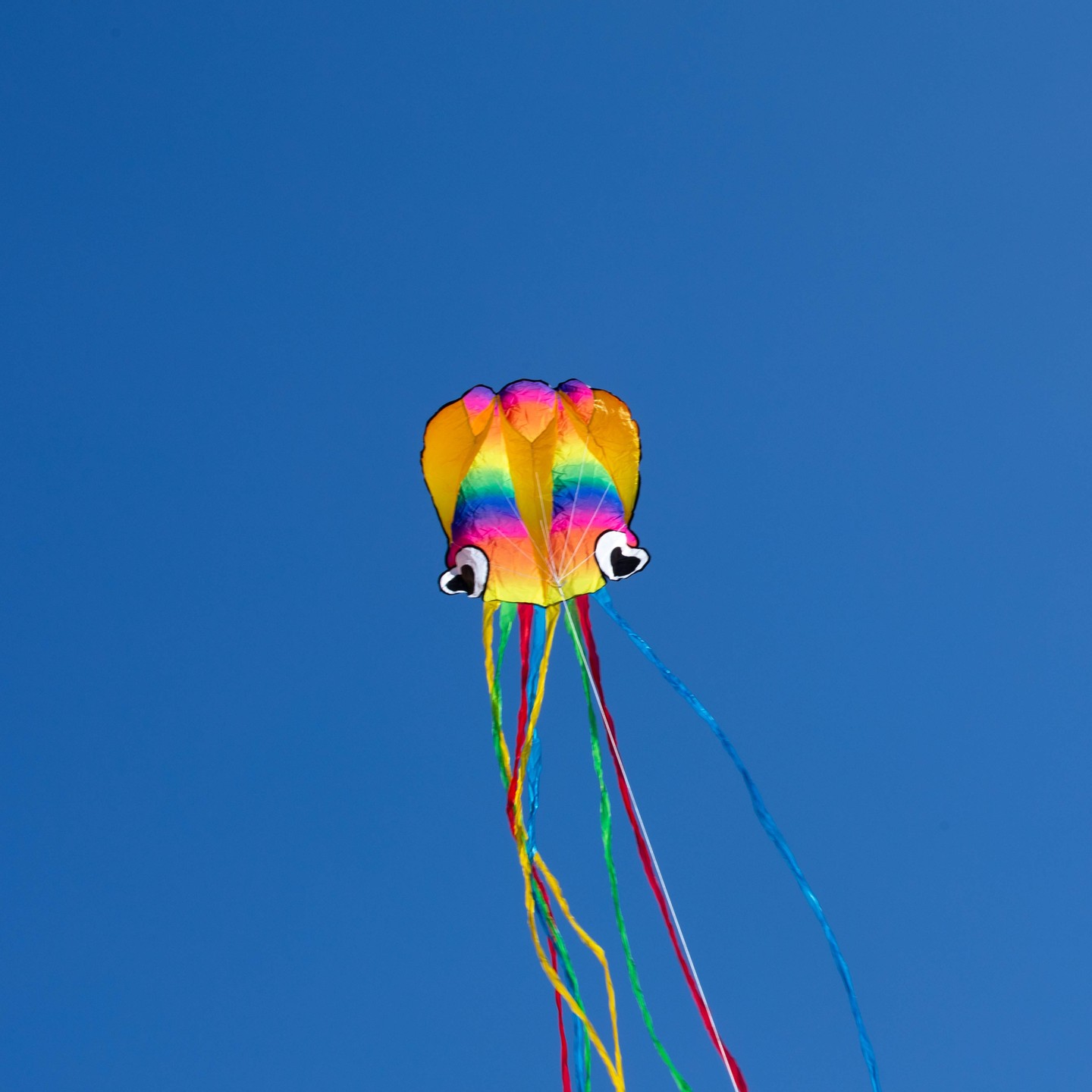 Colorful octopus-shaped kite with heart-shaped eyes flying against a clear blue sky, trailing multicolored ribbon tails.
