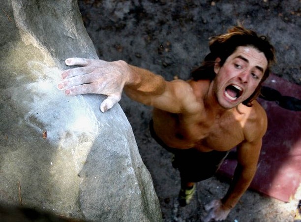 Shirtless man bouldering on a rock, gripping a hold with chalked hand, mouth open, straining upward with crash pad below.