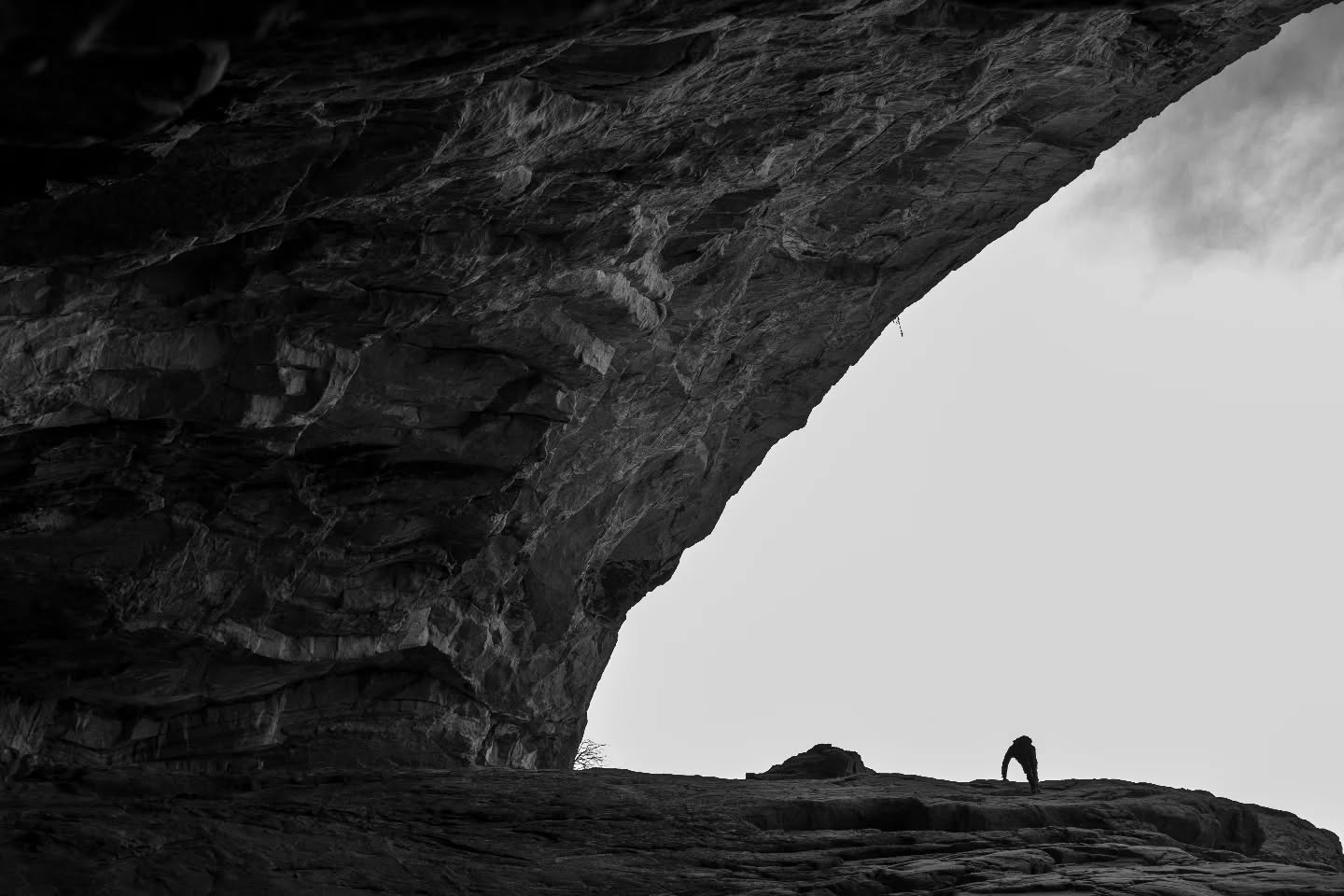 Black-and-white photo of a large rock arch overhang with a lone silhouetted figure standing on a ledge below it.