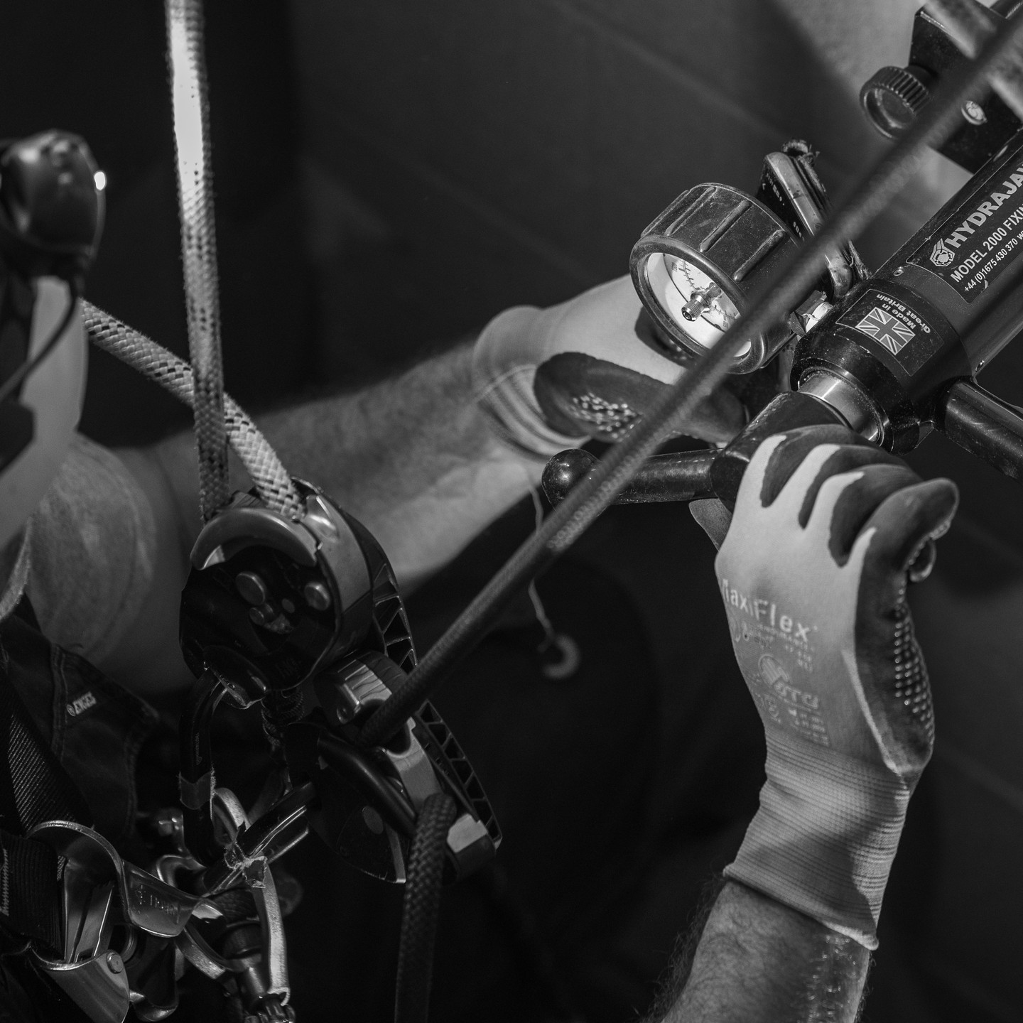 Black and white close-up of gloved hands operating a Hydrajaws tension gauge tool with rope and rigging hardware.