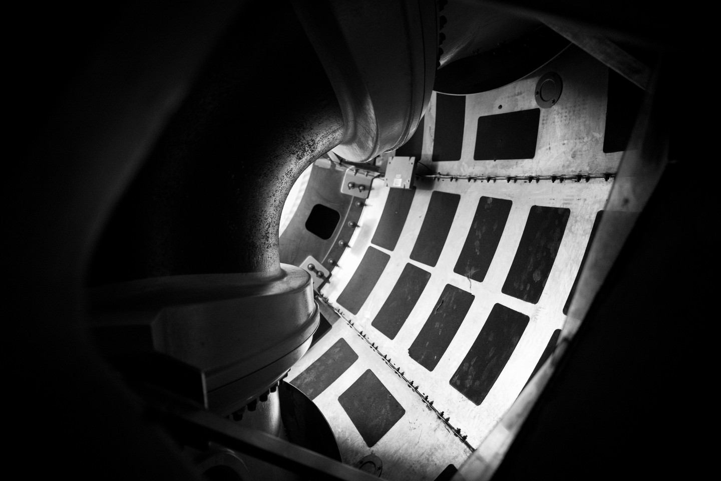 Black and white close-up of a curved metal duct opening beside a riveted panel with dark rectangular patches, industrial interior.