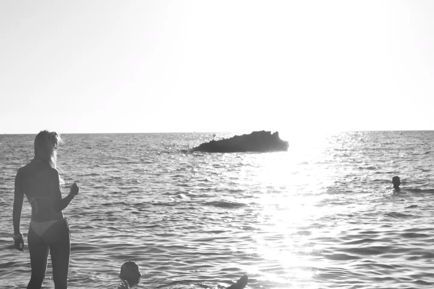 Black-and-white photo of a woman standing in the sea, swimmers nearby, sunlight reflecting on water, small rock formation in 