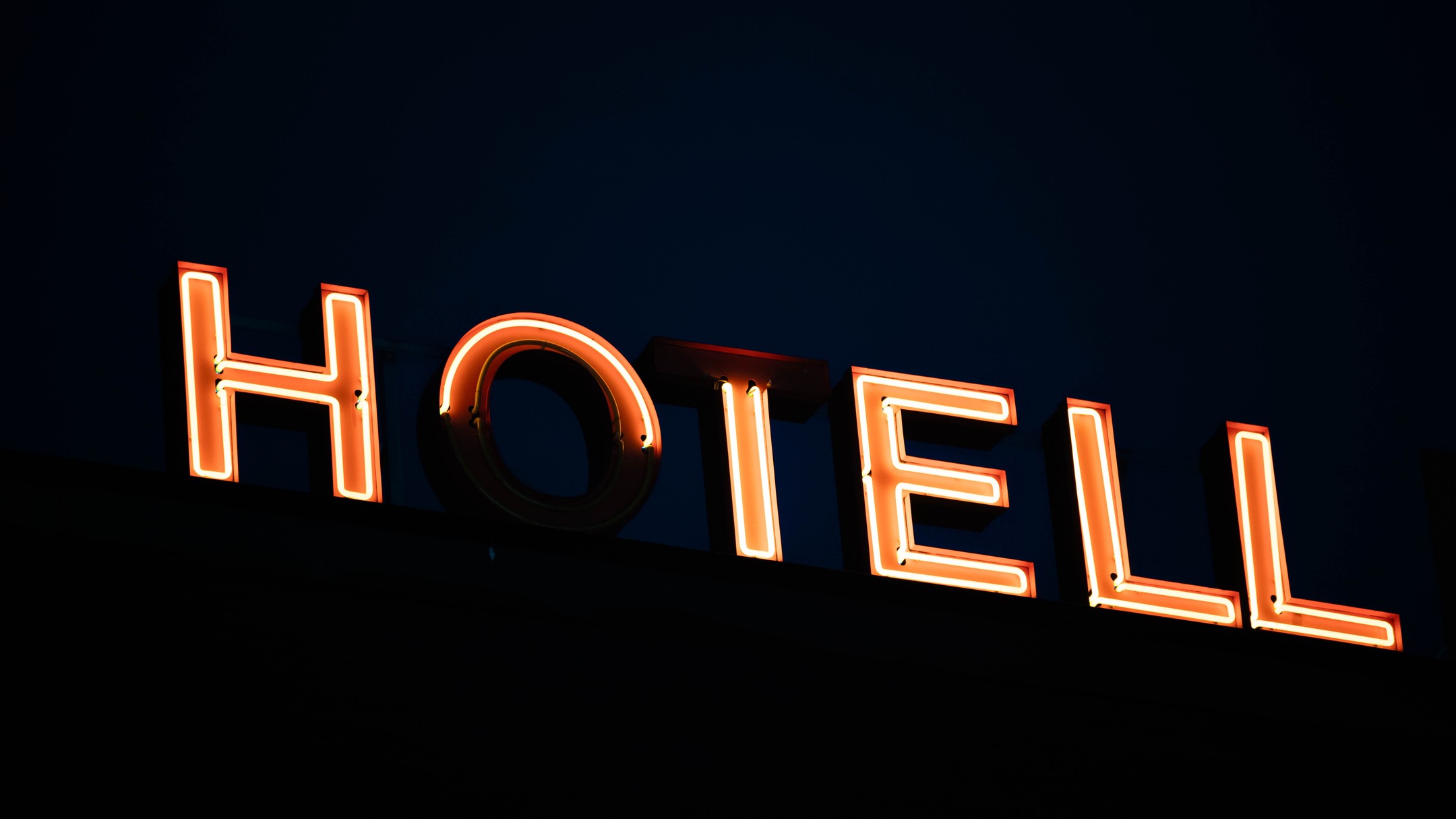Illuminated orange neon "HOTELL" sign mounted at an angle atop a dark building against a night sky.