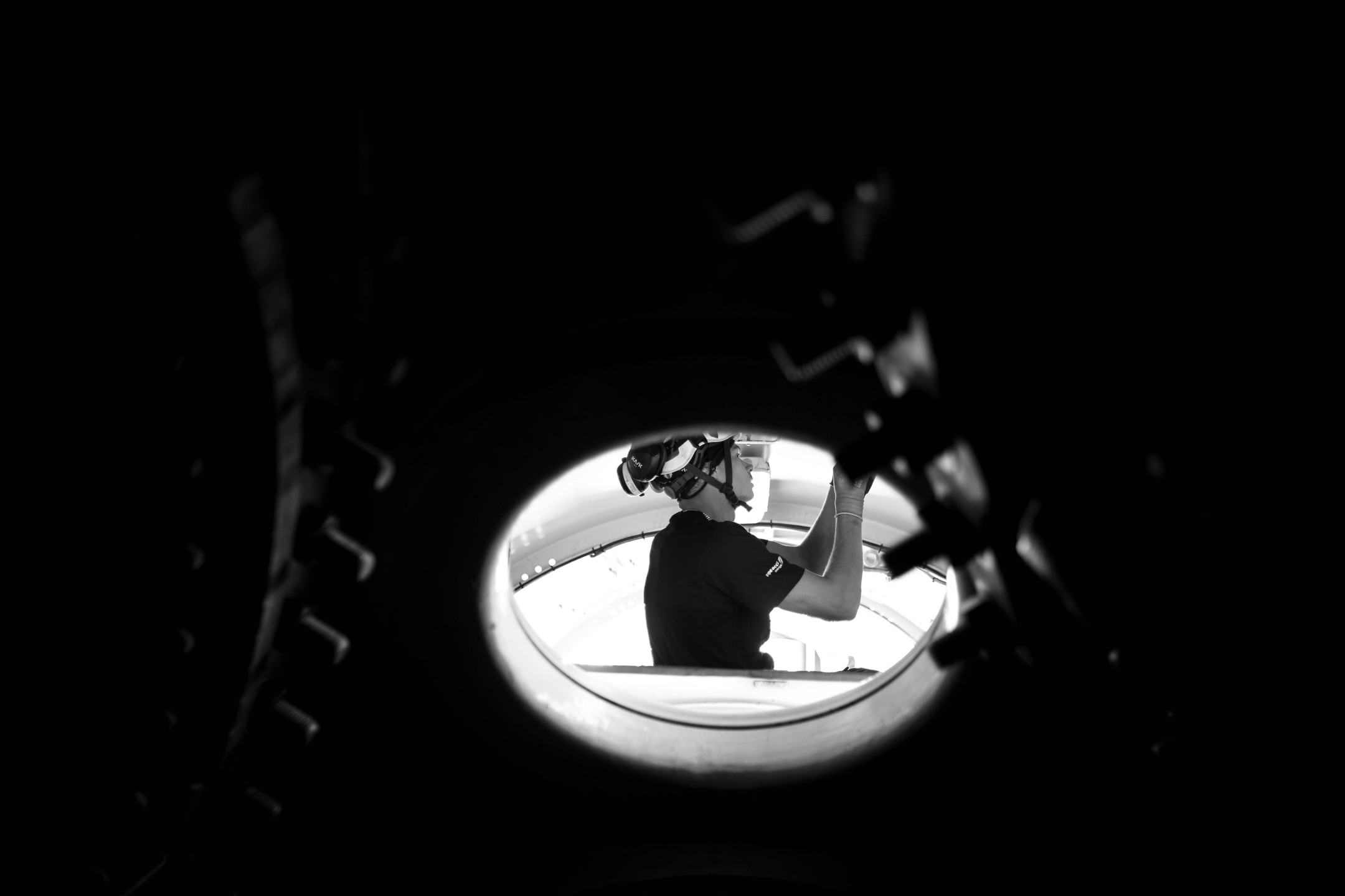 Black and white photo of a helmeted worker seen through a circular opening, reaching upward inside industrial machinery.