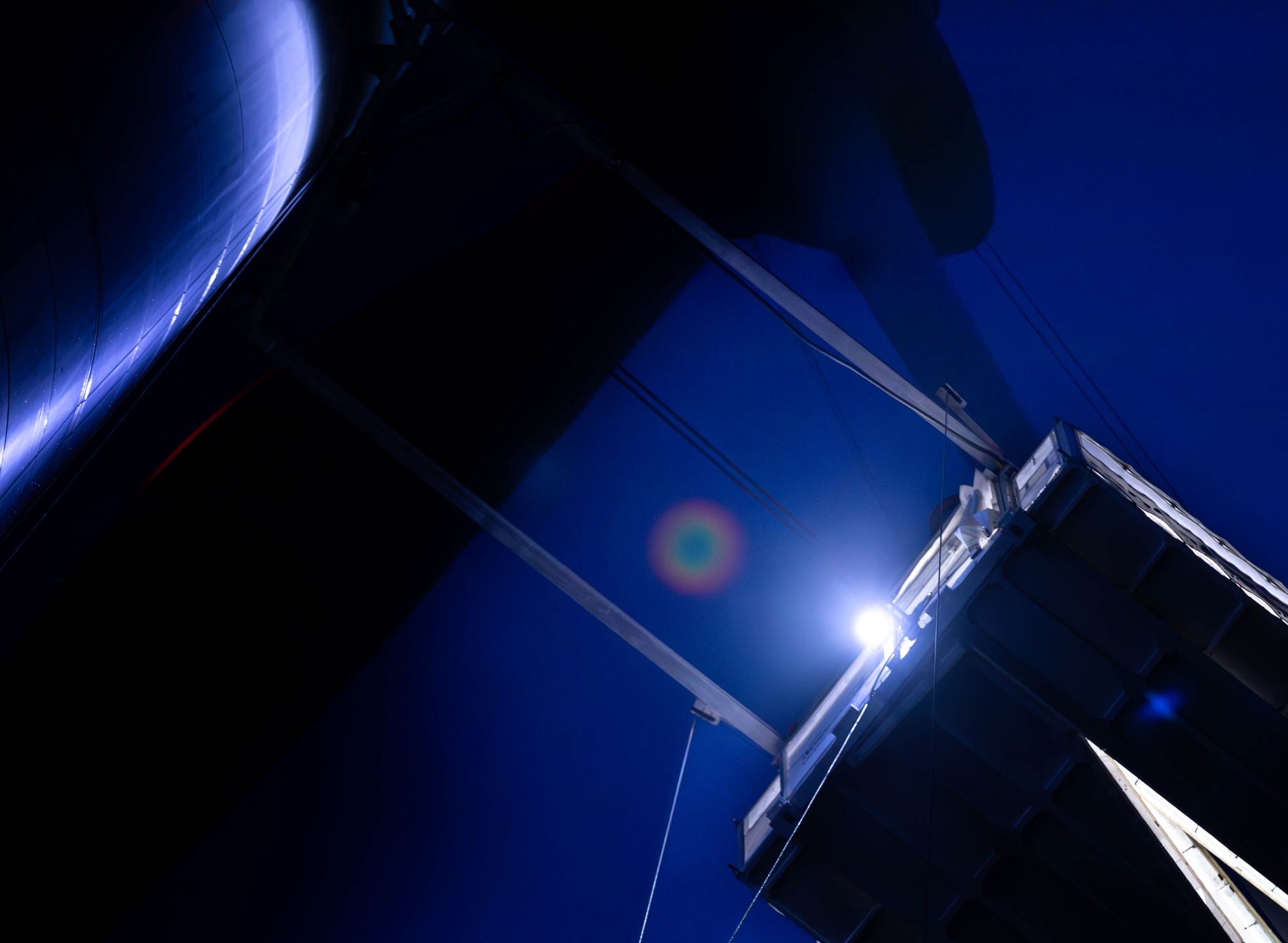 Upward view of wind turbine tower and blade against dark blue sky, with a bright light and lens flare.