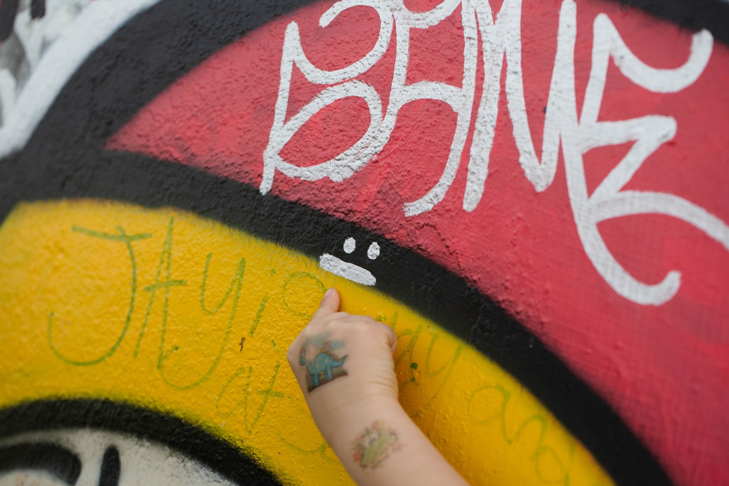 Close-up of colorful graffiti mural with white lettering on red and yellow background; a hand with small tattoos points near a painted smiley face.
