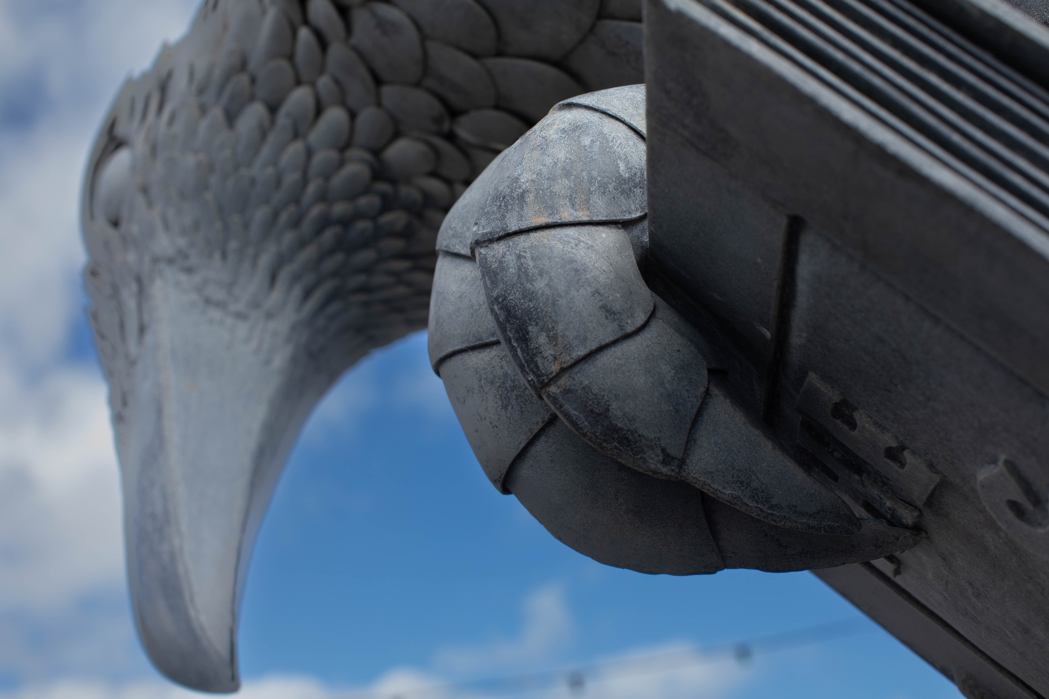 Close-up of a stylized metal eagle sculpture's head and curled wing against a blue sky with clouds.