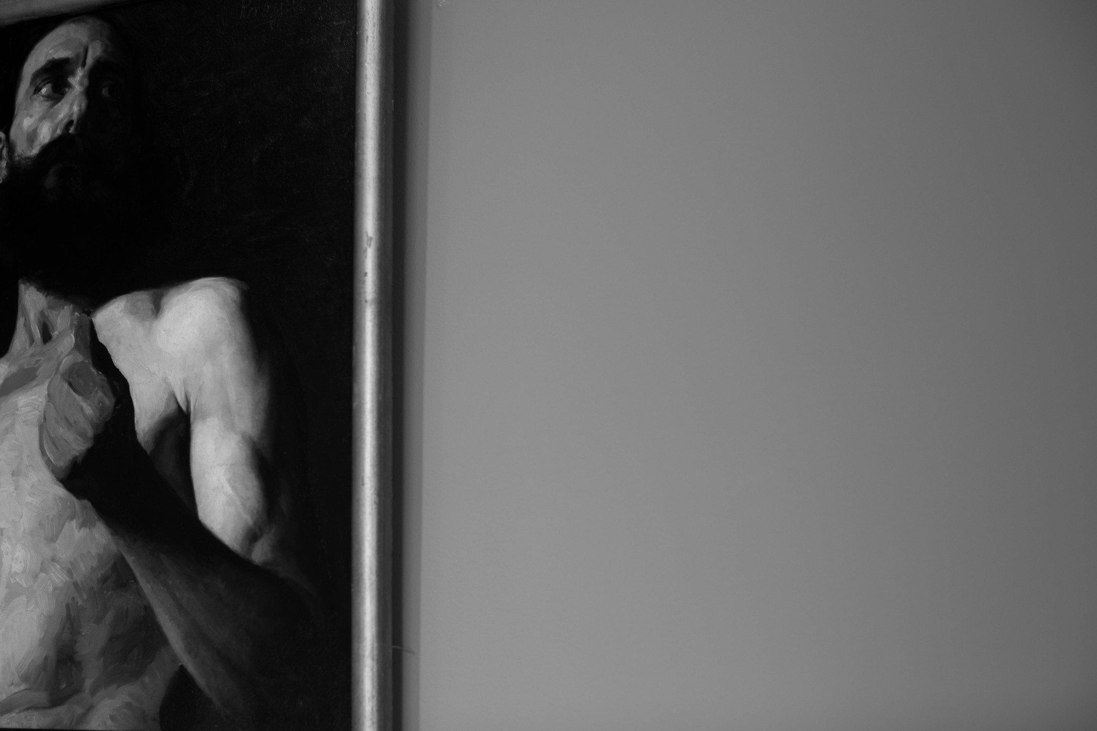 Black and white photo of a framed painting edge showing a bearded man's face and bare torso, against a blank gray wall.