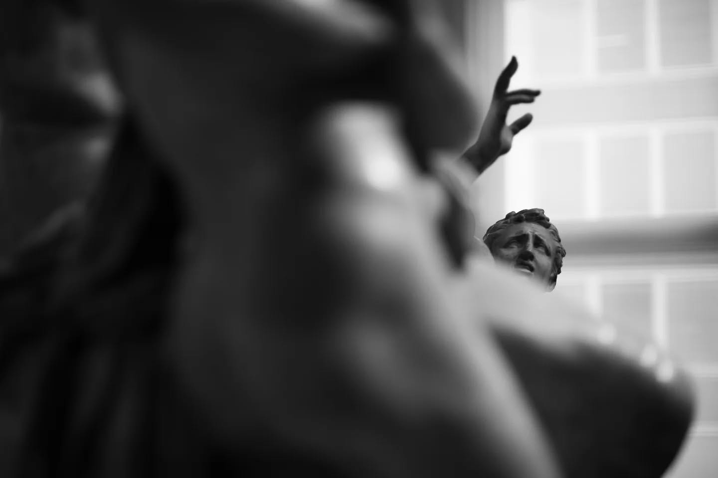 Black-and-white photo of a classical statue's head and raised hand, with a blurred figure in the foreground and a window behind.
