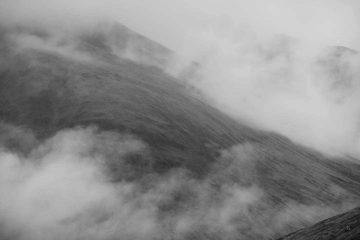 Black and white image of a mountain slope partially obscured by drifting clouds or mist.