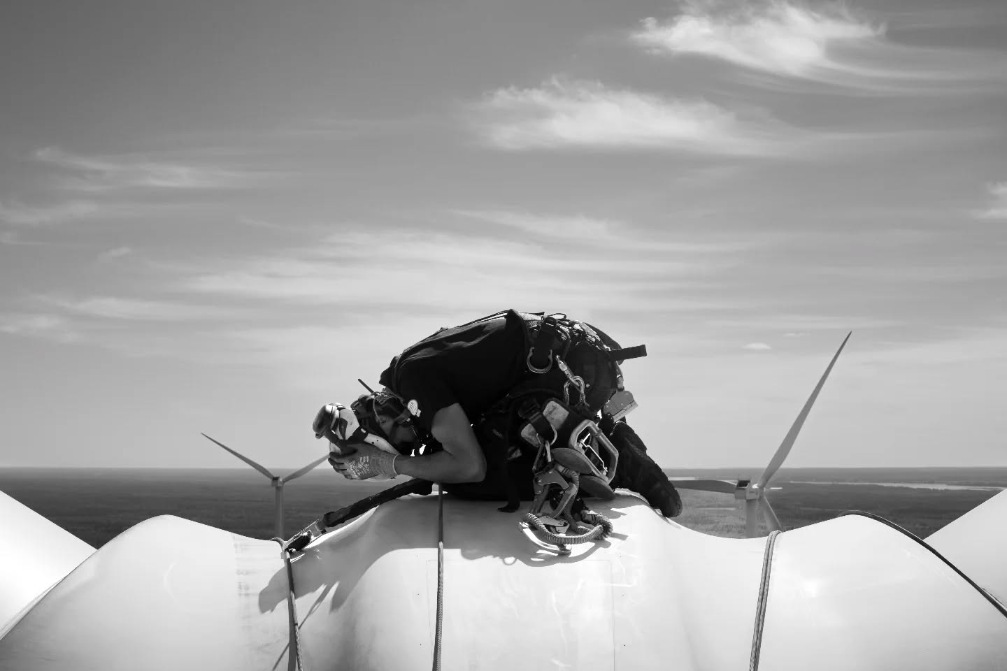 Black-and-white photo of a technician with climbing harness and rope gear lying atop a wind turbine blade, other turbines visible on the horizon.