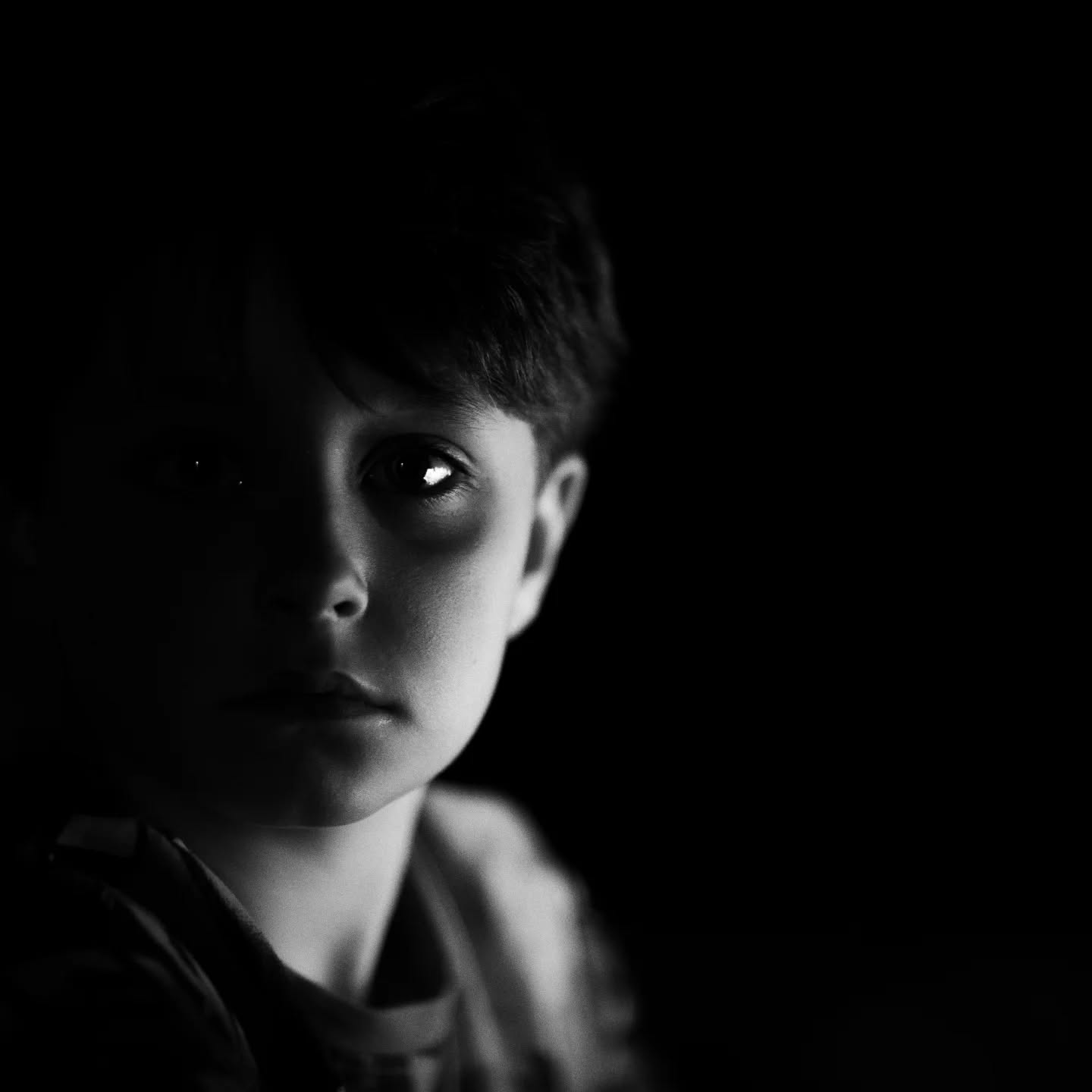 Black-and-white portrait of a child's face partially lit, one eye visible with catchlight, rest of face and background in shadow.