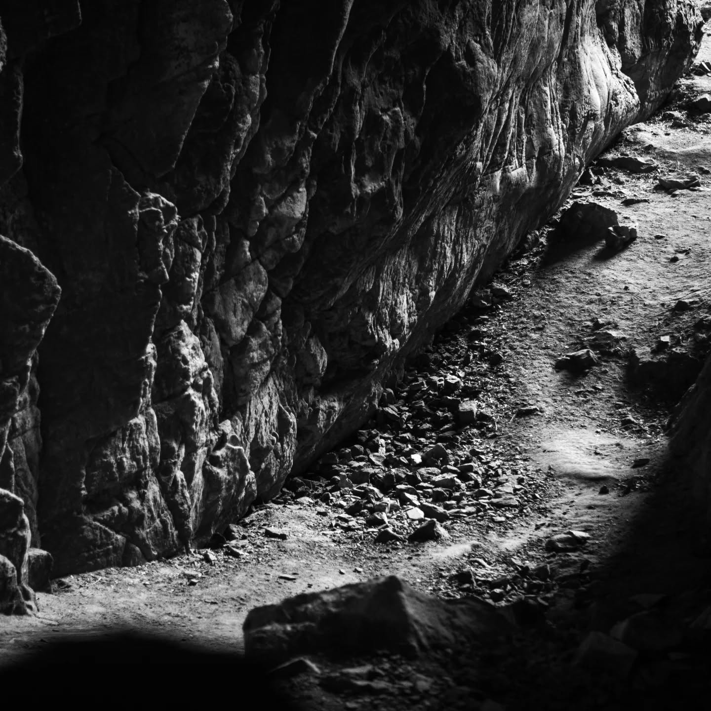 Black-and-white image of a rocky cave or gorge wall with a dirt path scattered with loose stones along its base.