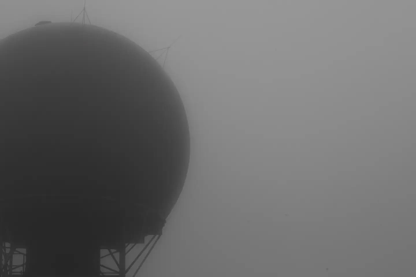 Silhouette of a large spherical radome atop a support tower, shrouded in dense fog, black and white.