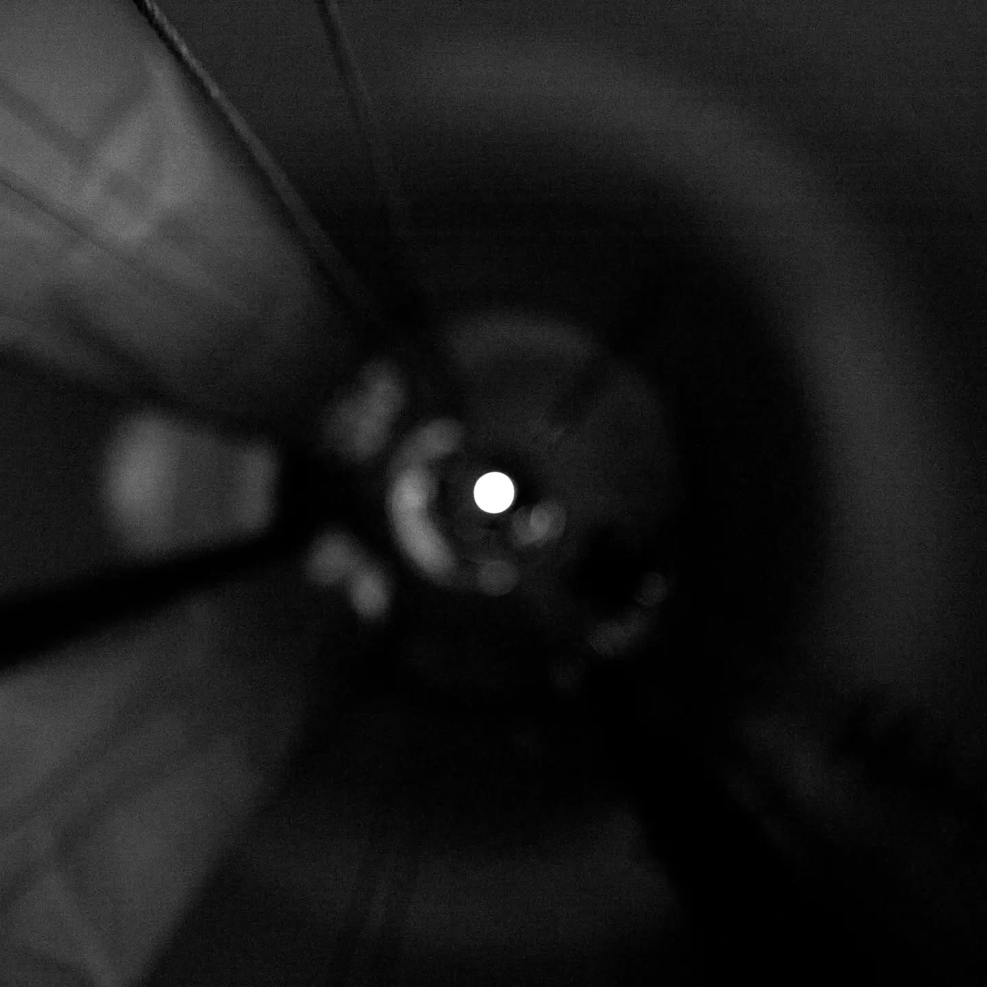 Black-and-white photo looking down a dark tunnel or stairwell toward a single bright light at the center.