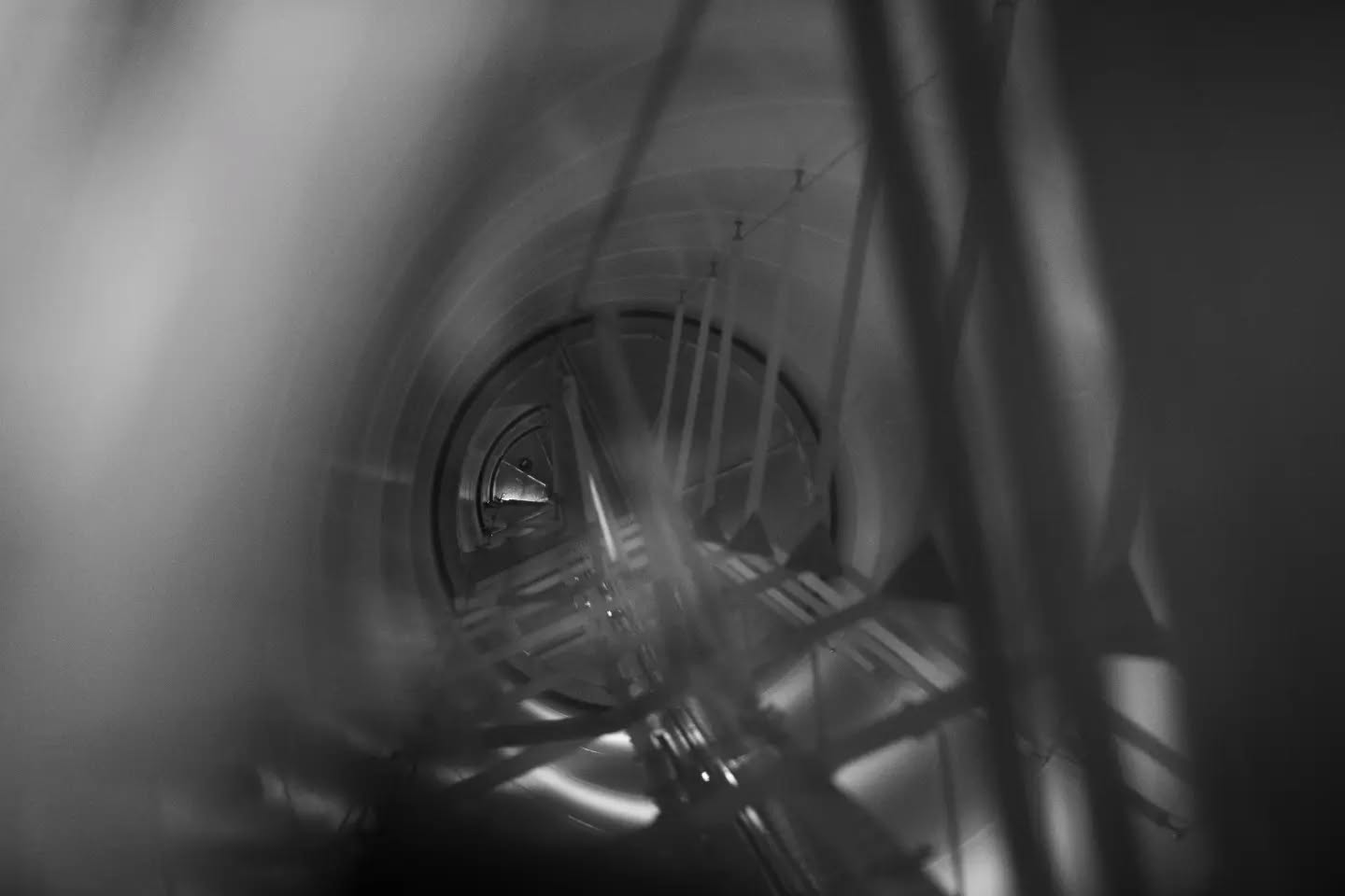 Black-and-white photo looking down a cylindrical shaft with metal rods and cross-bracing in the foreground, leading to a circular opening below.