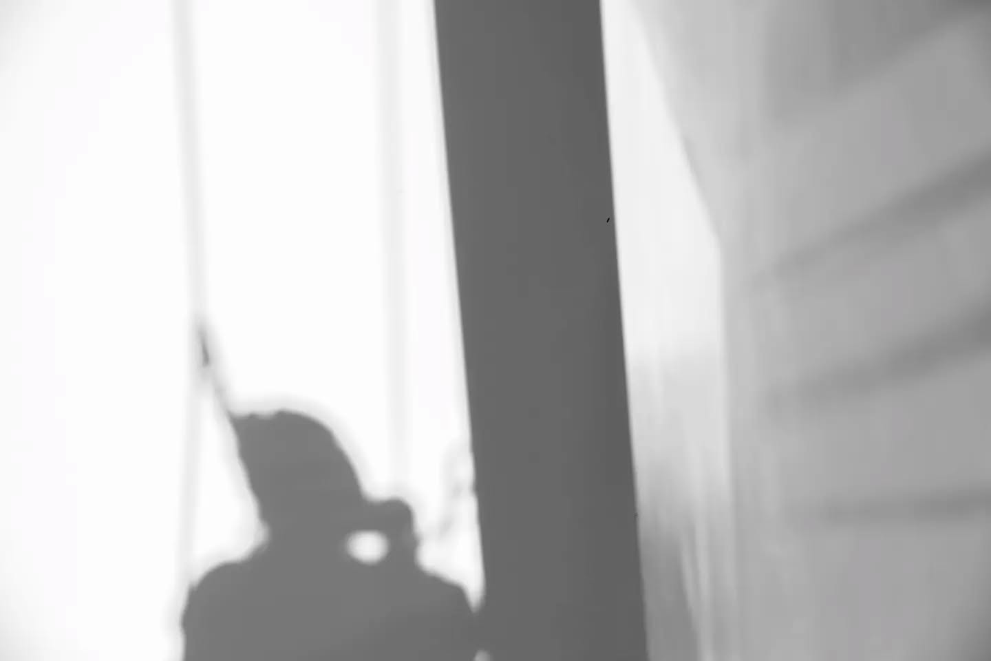 Black and white image of a shadow of a person's head and shoulders cast on a wall, beside a dark vertical architectural edge.