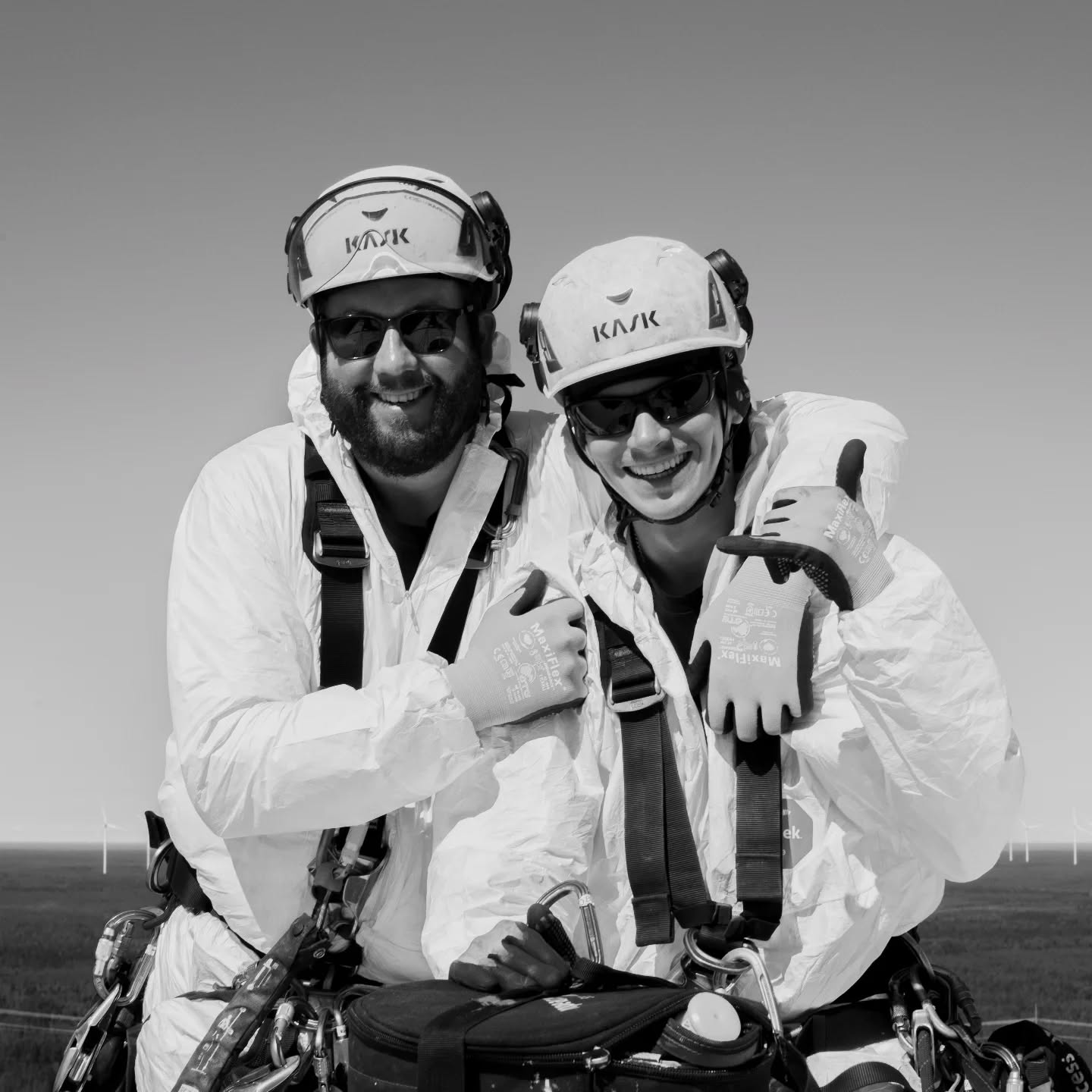 Black and white photo of two smiling workers in white protective coveralls, safety harnesses, KASK helmets, sunglasses, and gloves, giving thumbs up.