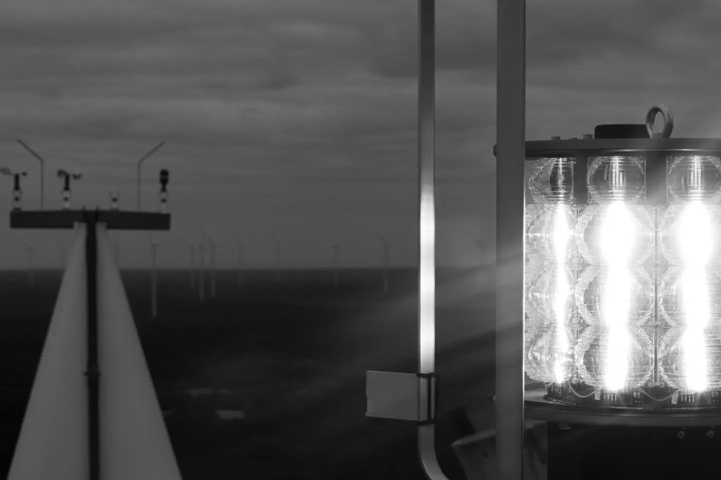 Black-and-white image of an aviation warning light with bright LED bulbs on a wind turbine, with turbine blade and offshore wind farm in background.