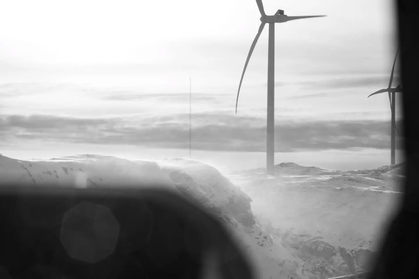 Black-and-white photo of wind turbines on a snowy ridge, viewed through a vehicle window with dark frame in foreground.