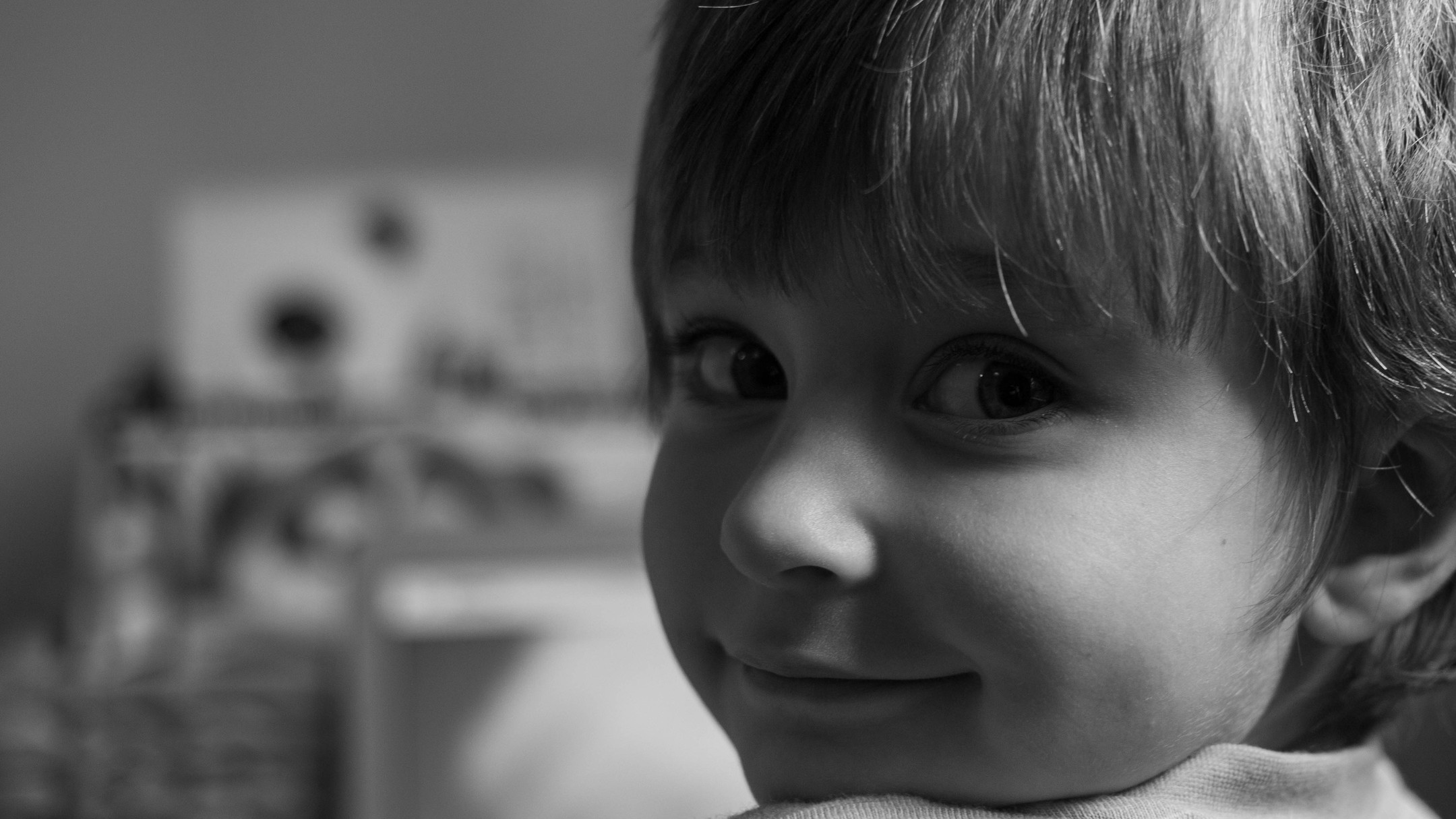 Black-and-white close-up portrait of a young child with bangs, looking sideways at the camera with a slight smile.