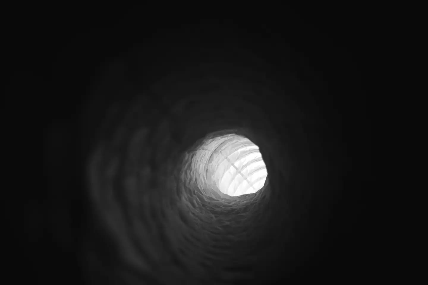 Black and white photo looking through a dark ribbed tunnel toward a bright circular opening with a grid pattern.