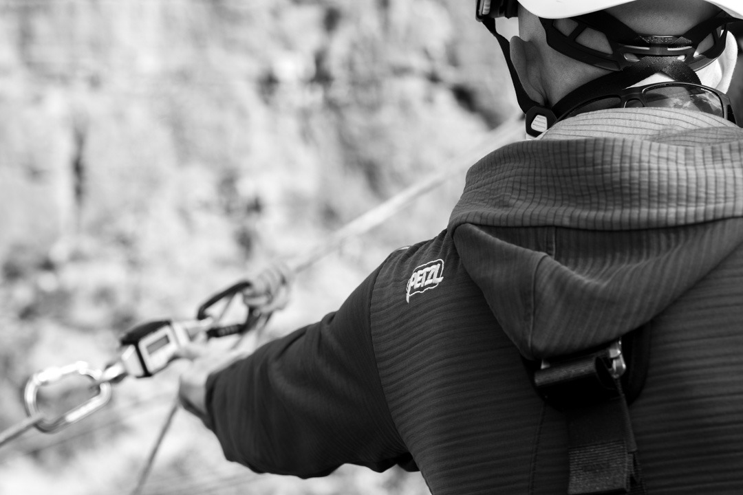 Black and white close-up of a person in a helmet and hooded jacket reaching toward a carabiner attached to a rope, cliff face blurred in background.