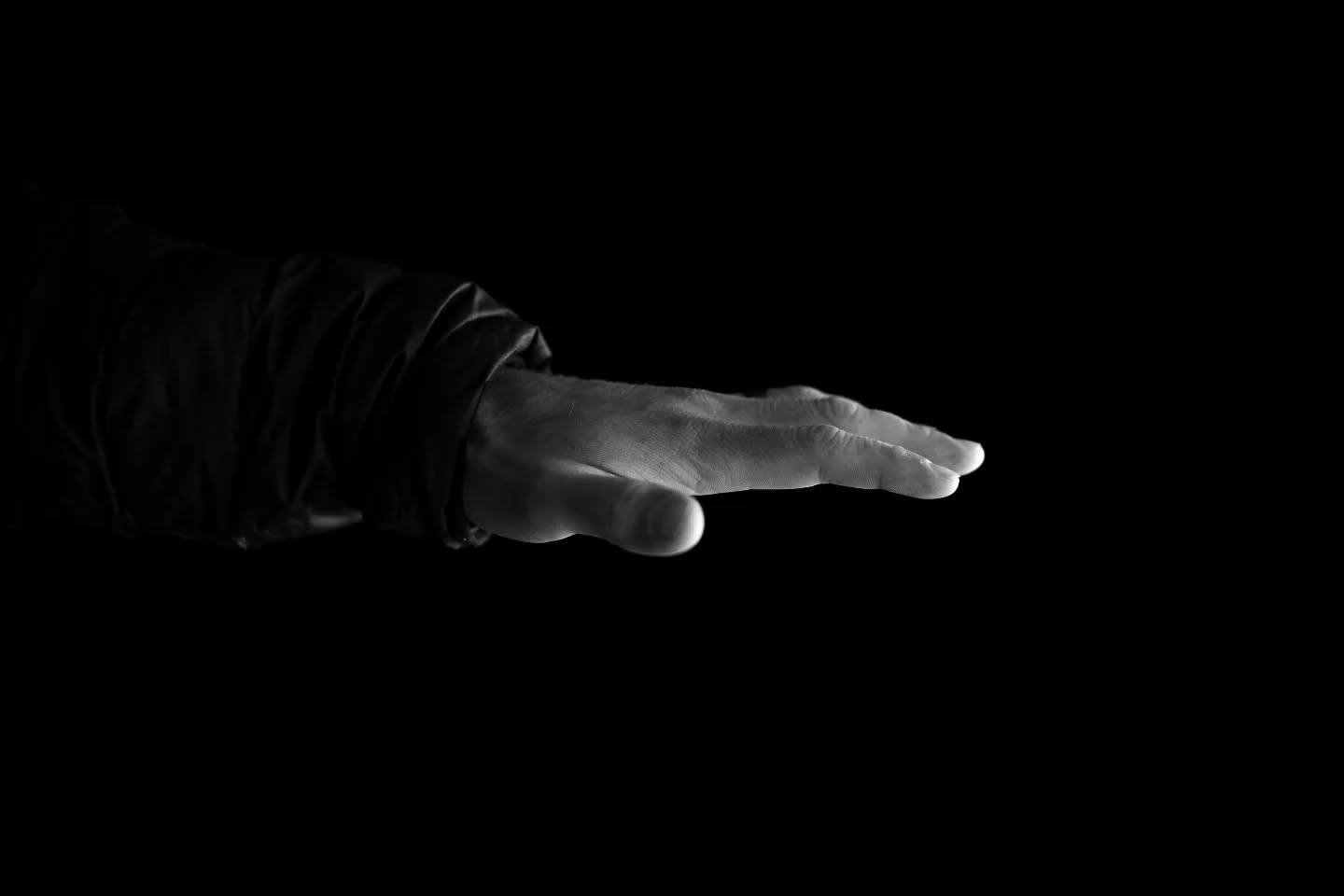 Black-and-white photo of an arm extending from a dark, rumpled sleeve, hand and fingers reaching out against a black background.