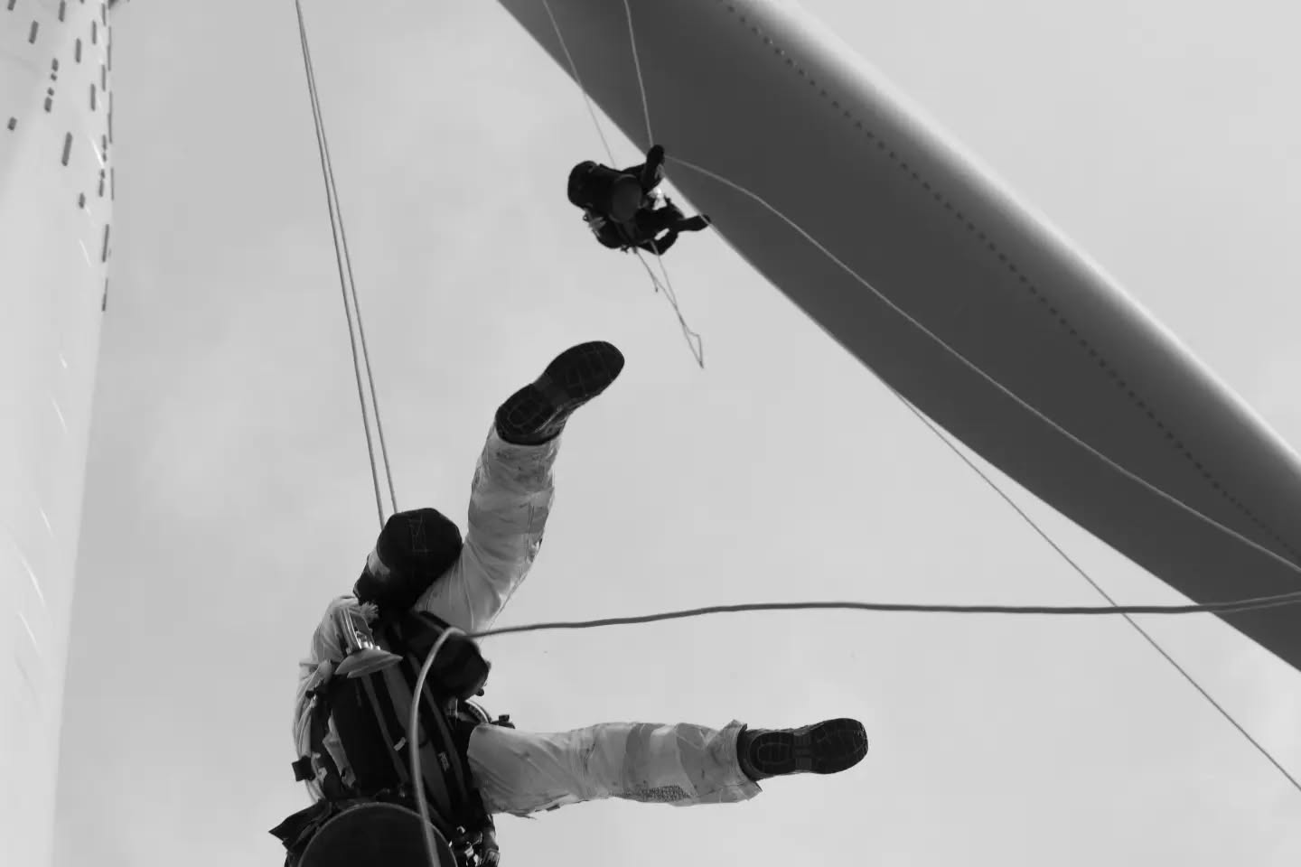 Black and white photo of two rope-access technicians rappelling near a wind turbine blade, viewed from below.