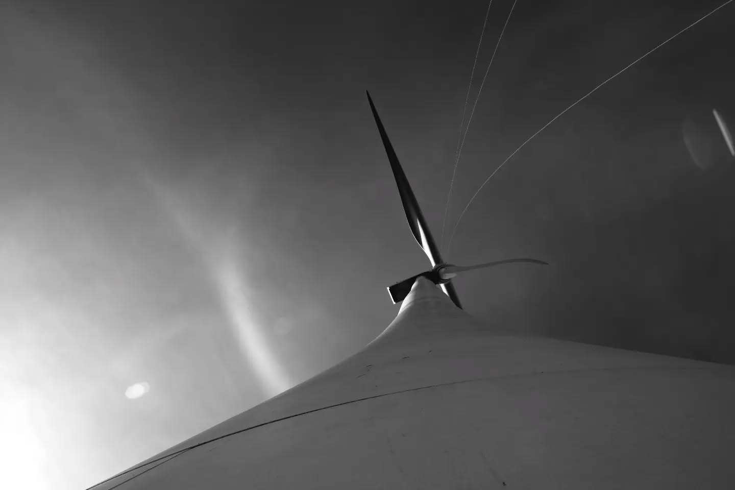 Black-and-white photo looking up at a wind turbine, blades motion-blurred against a hazy sky with faint light streaks.