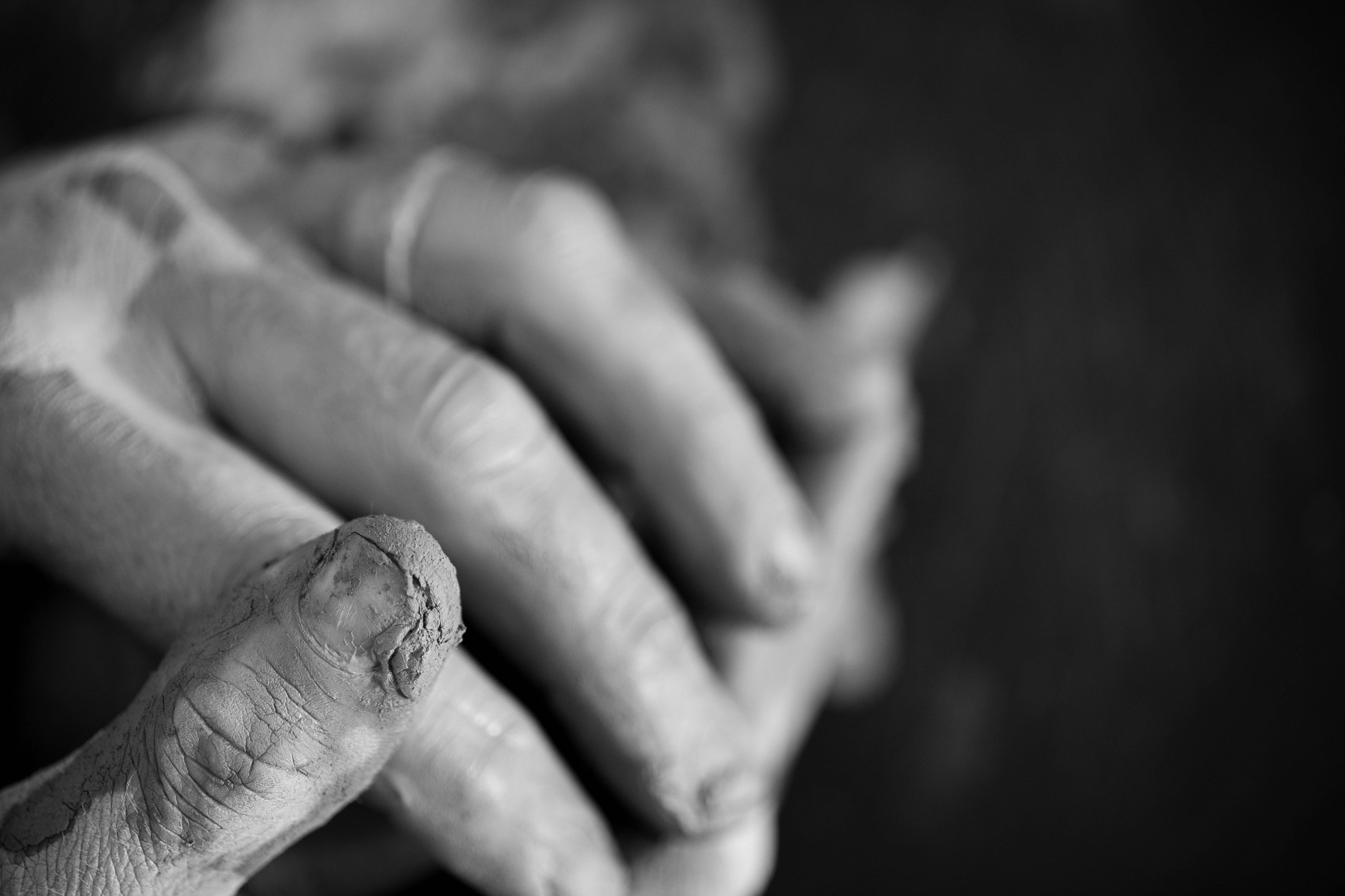 Black-and-white close-up of clasped hands with clay residue on the thumb, fingers interlaced, blurred background.