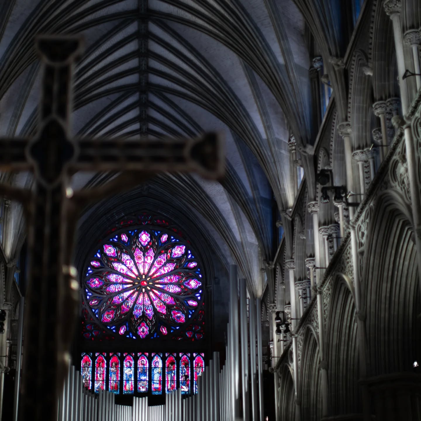 Cathedral interior with ribbed vault ceiling, rose window, organ pipes, and a wooden crucifix in foreground.