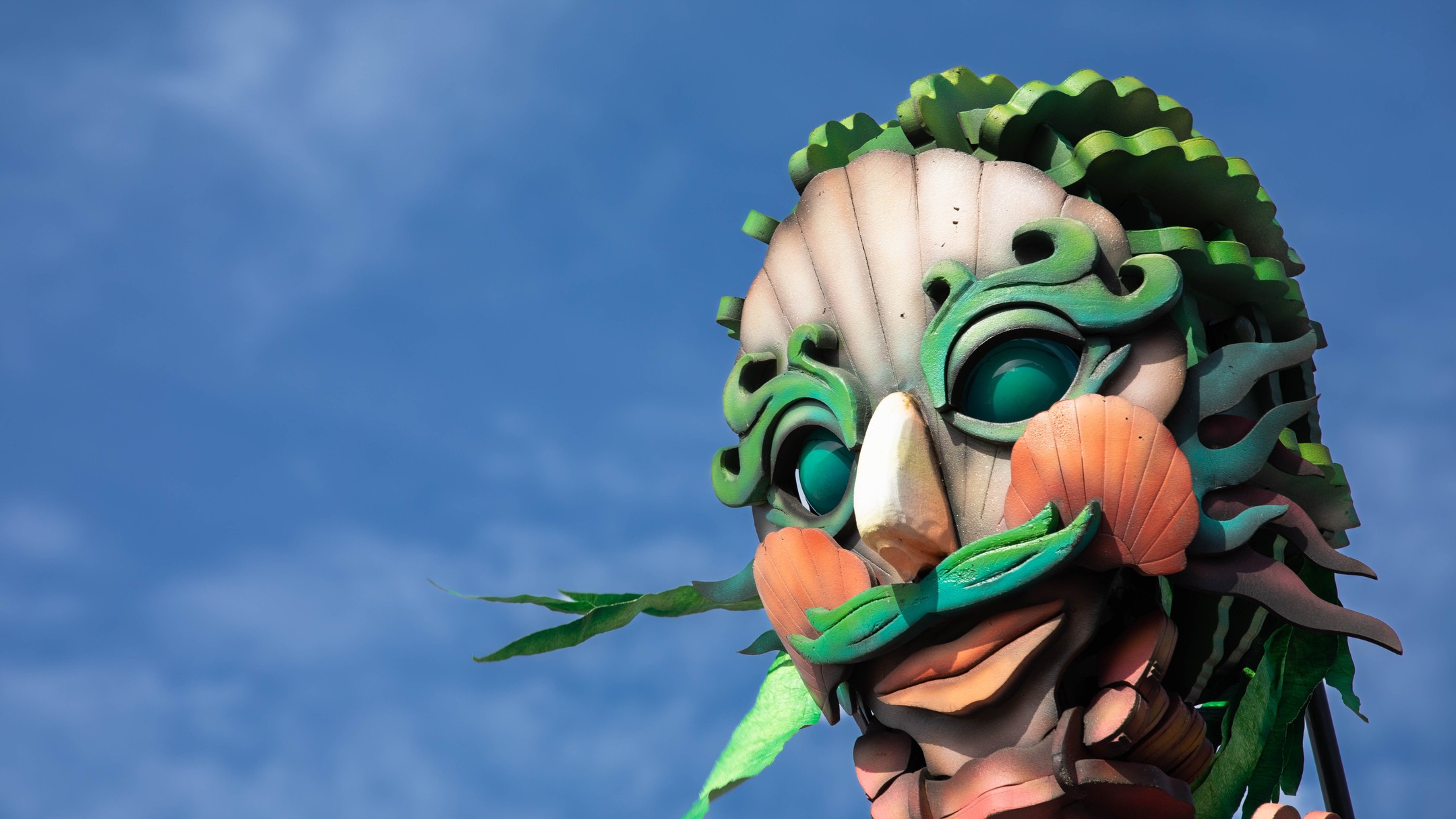 Close-up of a large sculptural puppet head with green swirled patterns, teal eyes, orange shell-like cheeks, and green leaf fringe against a blue sky.