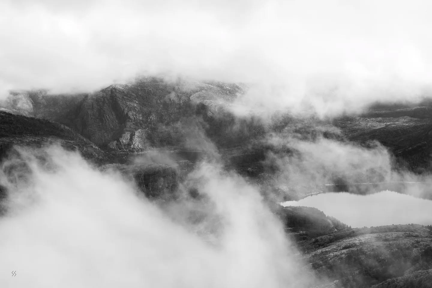 Black and white aerial view of misty mountains with a lake below, clouds drifting through rocky peaks and valleys.